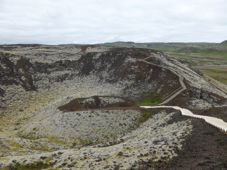 A dark red crater, covered in grey-green moss with a wooden boardwalk running across the rim and down into the middle.