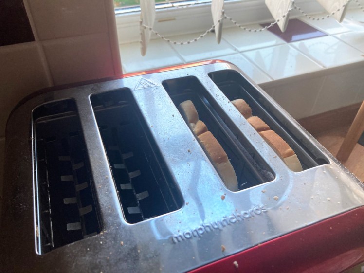 Two slices of toast in the right-hand slots of a four slot red toaster with a silver top. Behind you can see a tiled windowsill and off-white vertical blinds.