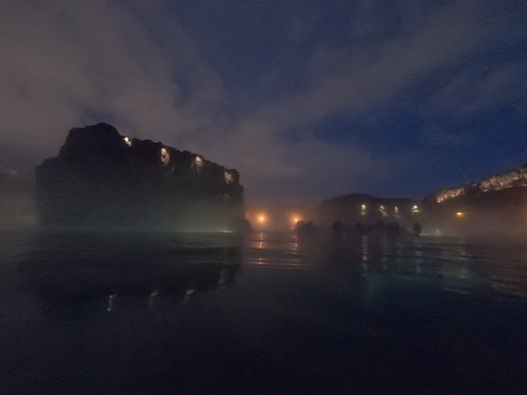 The Sky Lagoon by night. Cliffs rise out of water, with lights at the top and you can make out human silhouettes against the lights in the canyon. The