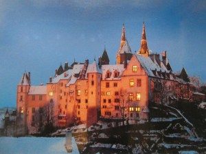 A postcard of Neuchatel Castle in winter at dusk. It has lots of turrets and spires and several of its windows are glowing.
