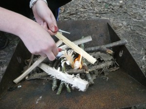 A wood fire being lit with a ferro rod in a rusty iron trough-shaped fire pit. There are three pieces of kindling cut into rough feather sticks and one that's been done well that's just starting to burn.