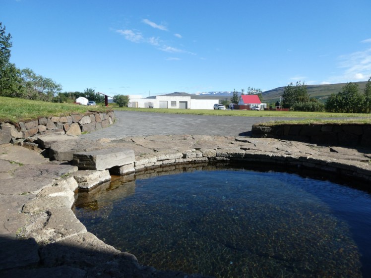 A shallow round pool surrounded by paving slabs. In the distance is a farm and a village.