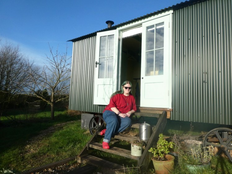 Me, sitting on the steps of a green shepherd's hut in the sun. I'm wearing a red t-shirt and blue jeans which are both big and baggy and unflattering, with red Crocs.