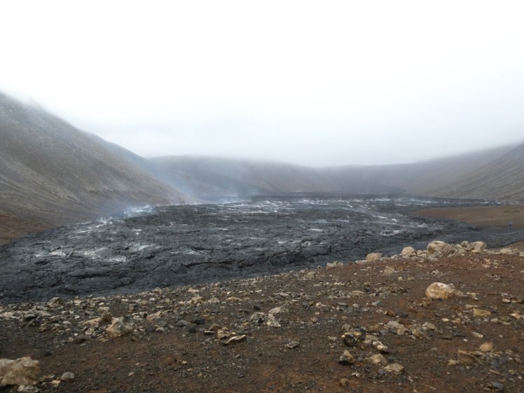 A thick layer of fresh black lava lies in a brown valley. The lava is flecked with white patches, as if it's turned to ash, and steam is wafting thickly from it in places.