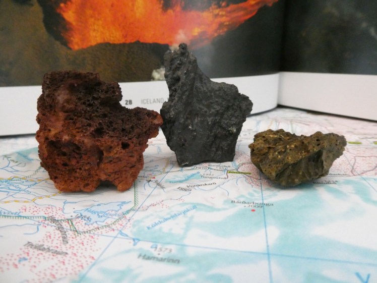 Three small pieces of volcanic rock on an Iceland map with a volcano book as a backdrop. From left to right, a reddish-brown piece of light crumbly rock, a sharp-edged dark grey piece of lava from the recent eruption, flecked with gold and silver, and a small piece of yellowish-green rock of similar light crumbly composition to the red one.