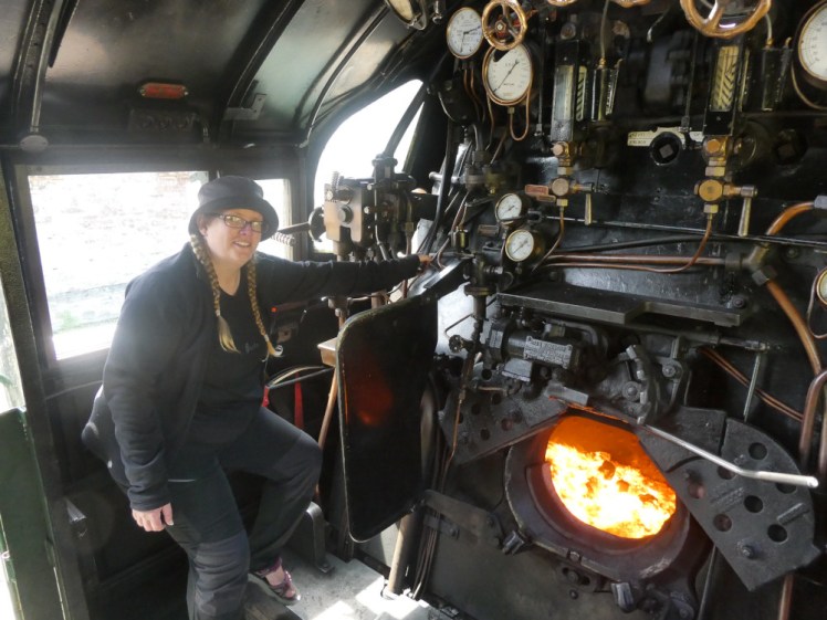 Me, dressed all in black except a navy bucket hat, sitting on the driver's seat of the steam loco cab, my hand on a bit of iron among a mass of iron levers, pipes and gauges. In front of me is the coal hole and it looks like the entrance to hell, a huge yellow-white fire.