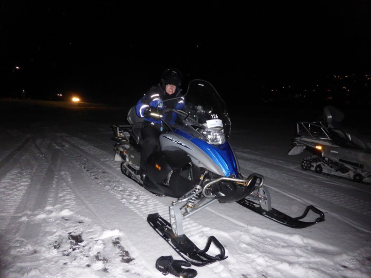 Me on a snowmobile in the dark. The blue snowmobile and I are illuminated only by the white light of the camera flash.