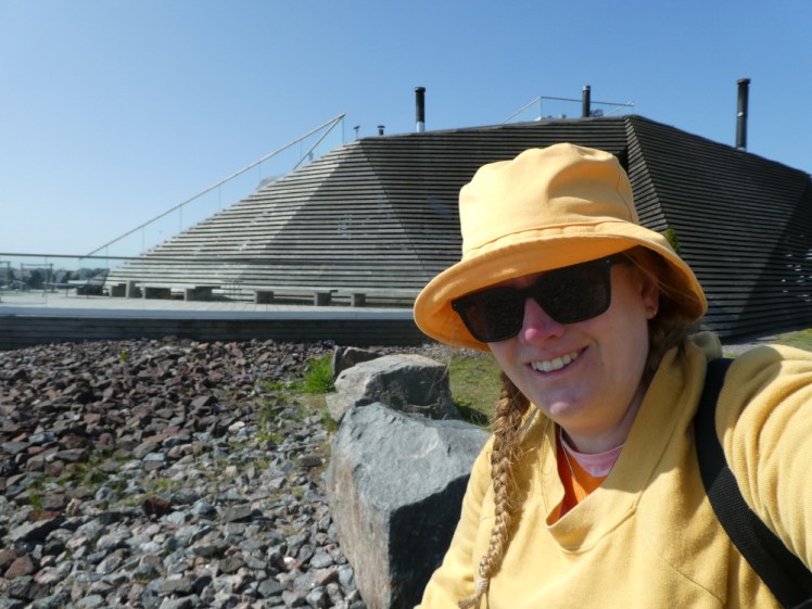A selfie outside Löyly. I'm wearing a yellow jumper and matching bucket hat with sunglasses. Behind me is an odd-shaped dome-like hive-like wooden building with a deck around it.