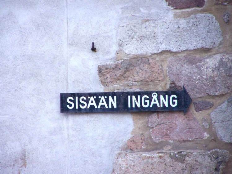 A black signpost on a partially-painted brick wall. It reads "entrance" in both Finnish and Swdish in nice clear white letters.