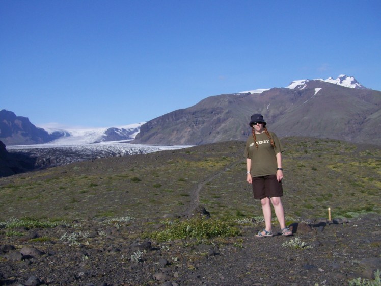 Another wonky timer selfie, this time of me in shorts in front of the glacial tongue Skaftafellsjokull. The sky is blue without a single cloud and the foreground is gravelly lava but with splatters of green and purple vegetation.