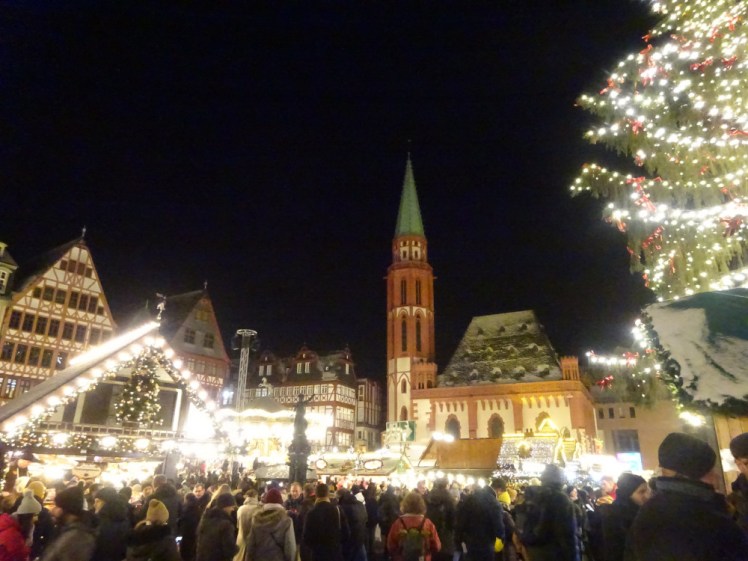 A Christmas market with illuminated stalls in a traditional Germanic square. In the background is a chuch with a reddish tower and a green spire. In the foreground is the edge of the huge Christmas tree.