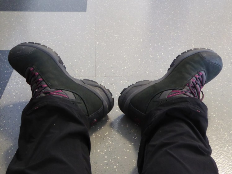 My feet splayed out on the floor of an airport, wearing fairly new hiking boots in dark grey with pink accents.