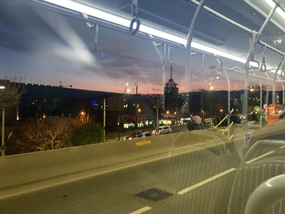 Through a bus window, complete with reflections, you can see a nearby mountainside with a pink & blue sunset over it and a tall building with a mesh tower and a light on top. Behind it, you can make out the lights of Tbilisi snaking up the mountain.