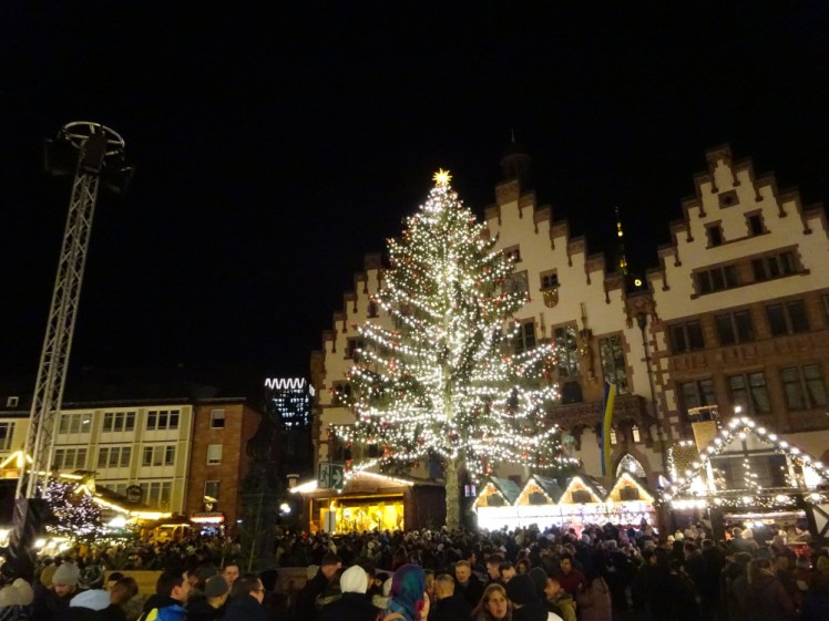 A tree has tall as a four or five-storey building lit with lights. Behind it is Frankfurt's Romer and below are various market stalls covered with lights and finally, there are hundreds of people crowded into the square.
