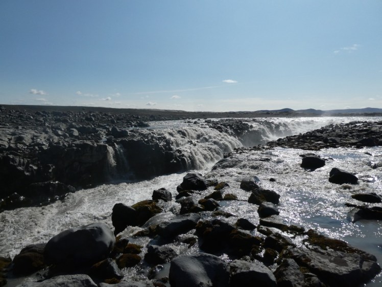 A shallow narrow black canyon filled with foaming grey water. You can see the river falling into the canyon as a waterfall on the other side. On the side near the camera, you can see how the water has wandered around near the top of the canyon, rather than staying neatly in any banks.