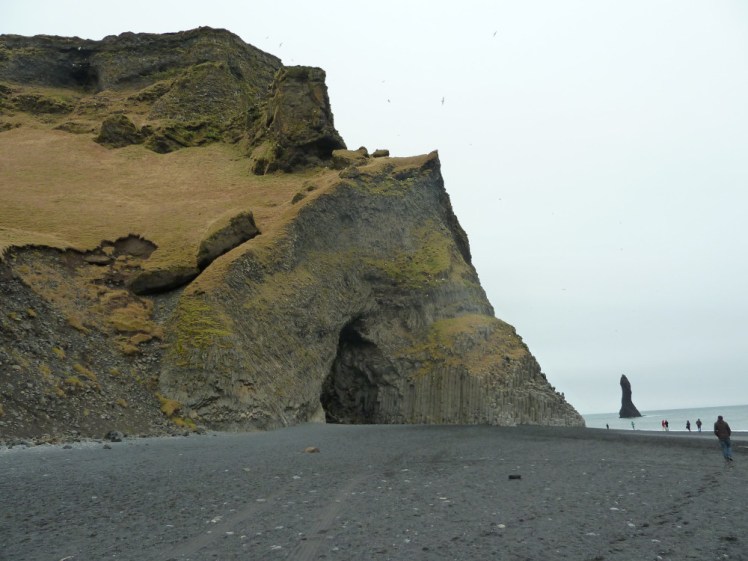 The headland at Reynisfjara, a big cliff covered in yellowish grass. The beach side rises up into a triangular rock face made of basalt columns and with a large cave mouth in it. In front is a black sand beach and to the right, the most violent and terrifying bit of sea in Europe.