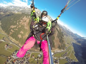 A paragliding selfie taken from a GoPro on a long selfie stick with remote control. I'm wearing pink trousers and red goggles and hanging in mid-air above a town and a mountain range. Behind me is my pilot, in a green jacket, holding onto the ropes of the canopy that's keeping us at this height.