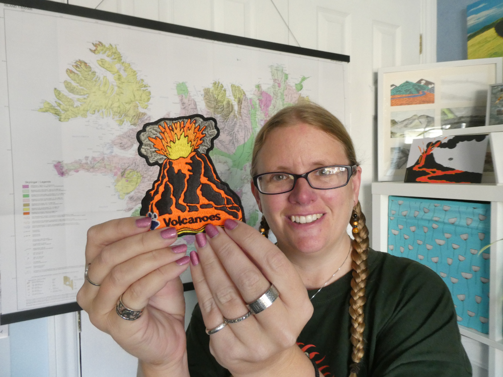 A selfie with plaits and a green t-shirt, holding up a large cloth badge with a volcano on it. I'm wearing lots of silver rings. Behind me is a tectonic map of Iceland and on the shelf behind me is a cartoon of a visitor to a volcano.