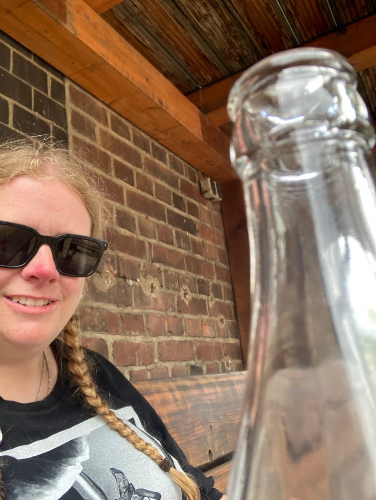 A selfie, with sunglasses on, sitting on a wooden bench with the underside of a wooden deck visible above me, against the brick wall of a warehouse. I've positioned the camera so it gets the neck of a glass bottle in the foreground.