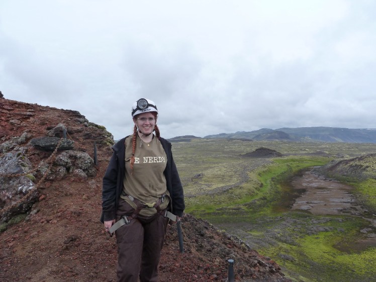 Me, in harness and helmet, standing on top of a small volcano with a green-grey lava field behind me.