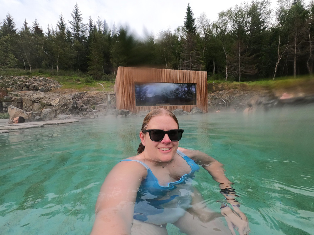 A selfie in the Forest Lagoon, wearing a swimsuit with a polar bear on it. The water is jade green, there's a wooden rectangular building behind me and the whole thing is set in front of a birch and pine forest.