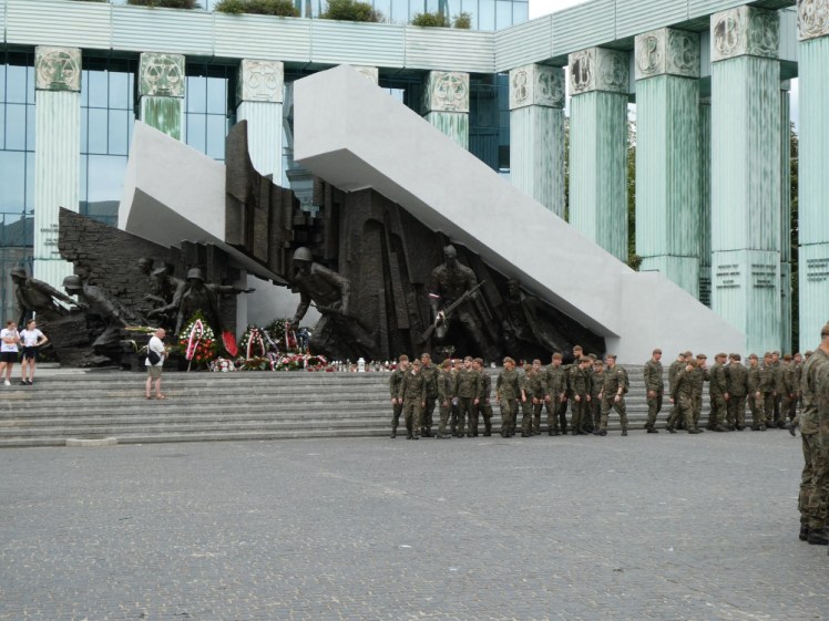 A ceremony with soldiers in camouflage standing by the Warsaw Uprising Memorial.