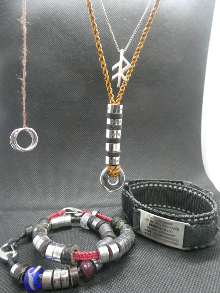 A pair of tiny silver hoop earrings hanging from a brown thread; a silver rune pendant hanging from its chain with a necklace made of twisted brown threads and threaded with stainless steel circles engraved with various things. Lying underneath are two bracelets in the same style, with more "steps" on them, and a webbing bracelet with reflective thread around the top and bottom. The plate giving my emergency contact details has mostly been blurred out.