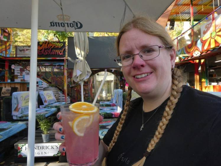 A selfie at a pop-up cafe made mostly out of shipping containers and pallets. I'm holding up a glass of pink melon lemonade with slices of orange in it.