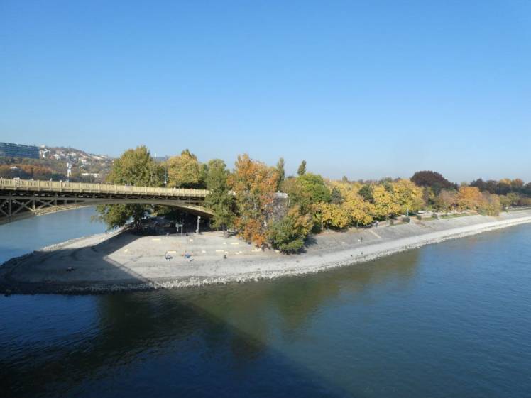 A view of Margaret Island from the bridge, a tree-covered island with concrete sides, connected to the bridge across the Danube by another bridge.