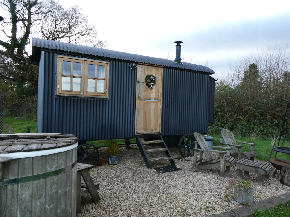Outside the Happy Hare, a dark grey-blue shepherds hut with wooden door and windows. In the foreground is the side of a more conventional hot tub, with wooden sides and a plastic lining.