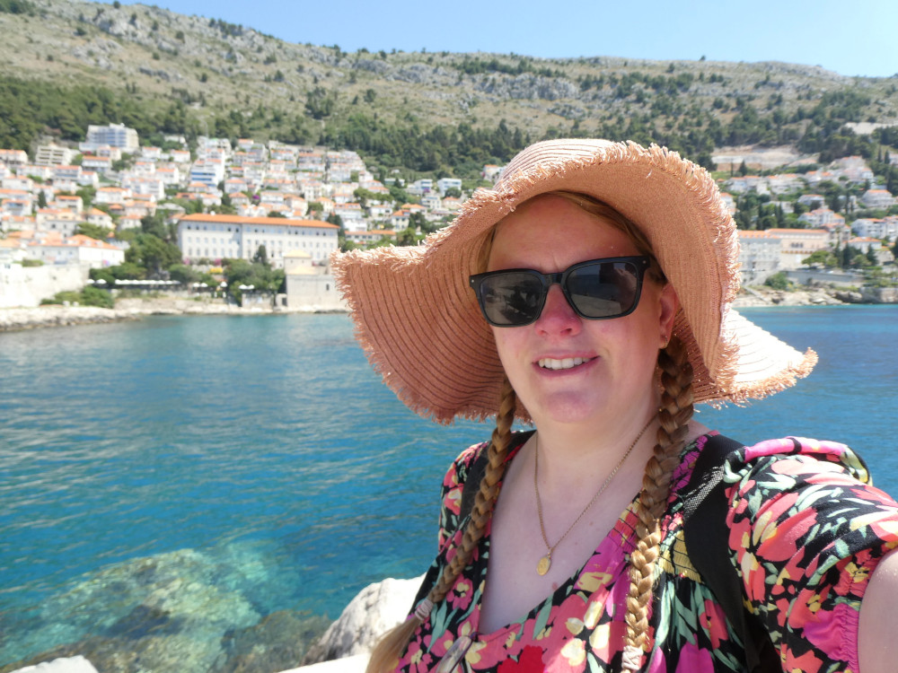 A selfie on the waterfront in Dubrovik with the town on the hillside behind me and some very blue water. I'm wearing a pink flowery dress and a very big floppy pink straw hat.