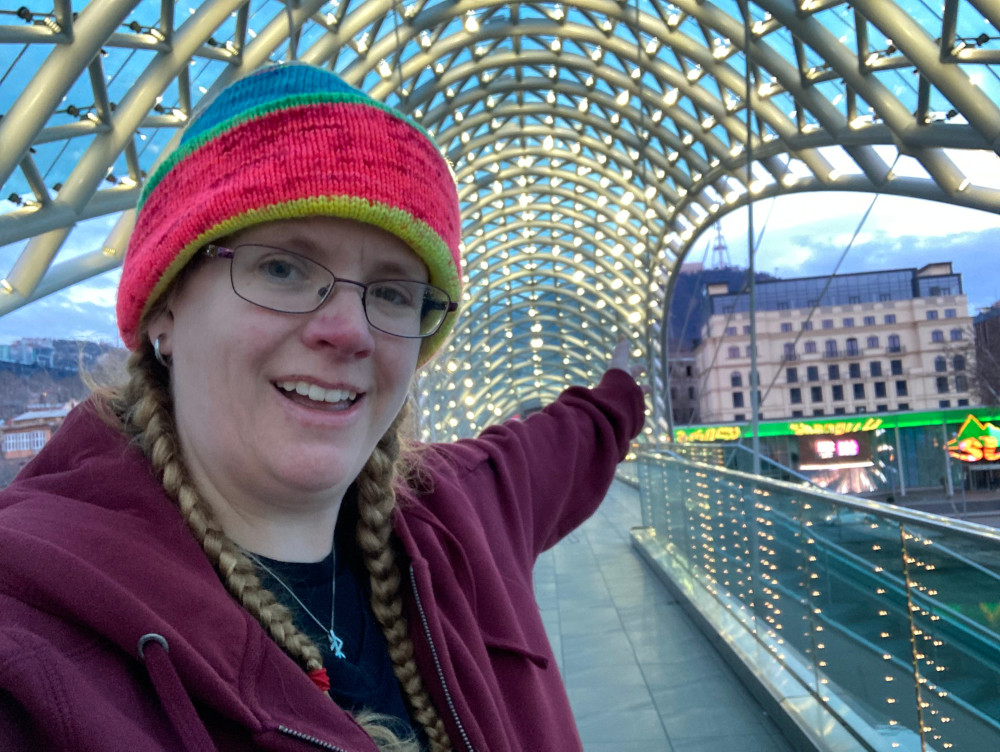 A selfie, in a burgundy hoodie and colourful hat, standing on the Peace Bridge, which is a sort of curvy glass bridge inlaid with thousands of little lights, just early enough in the morning that you can still see the lights.