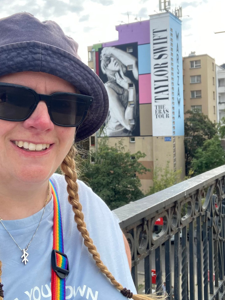A selfie in Warsaw in a pale blue t-shirt and a navy bucket hat in front of a Taylor Swift mural on the side of an apartment building.