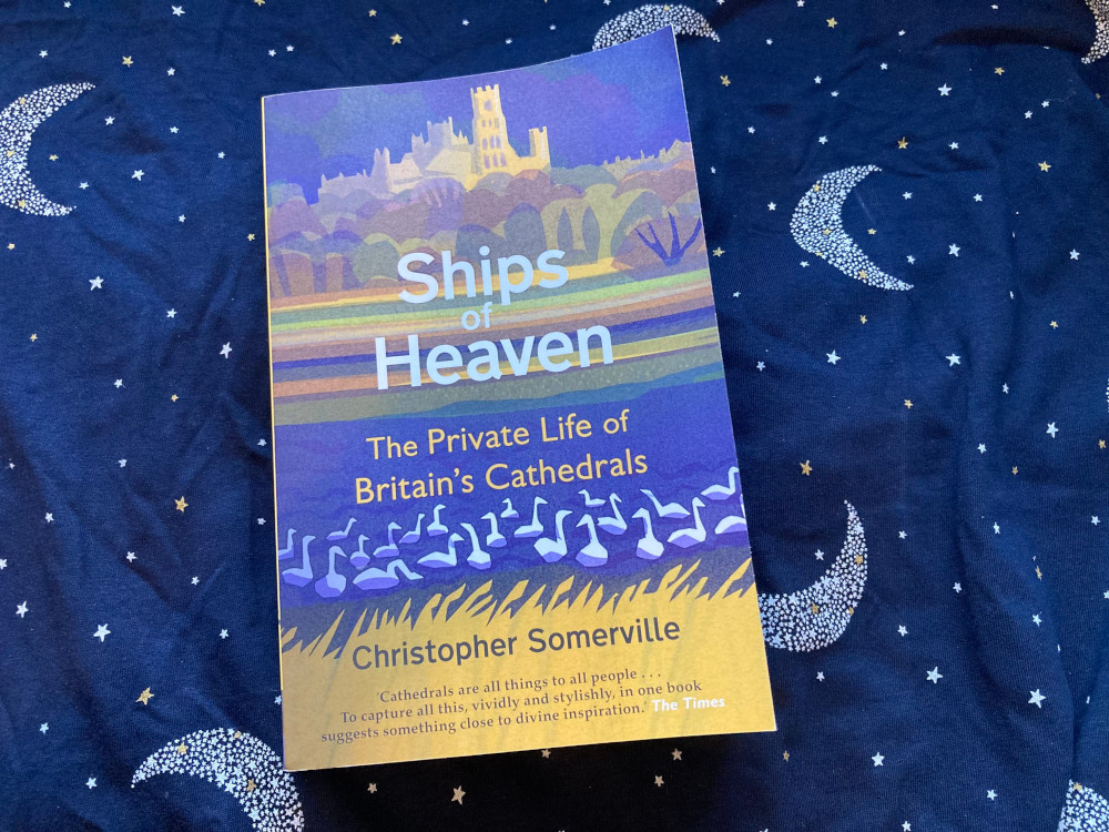 Ships of Heaven by Christopher Somerville, a paperback book with a view across a field towards a cathedral in shades of blue and yellow.
