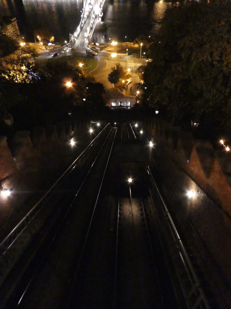 The view from the front seat of the funicular, looking down the sloped tracks towards the valley station and the road and the start of the Chain Bridge, most of it lit in yellow because it's night time but the bridge is lit in white.