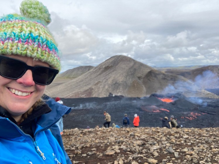 A selfie with a very small volcano behind me. There's orange lava bubbling in the crater and a cloud of blueish smoke coming off it.