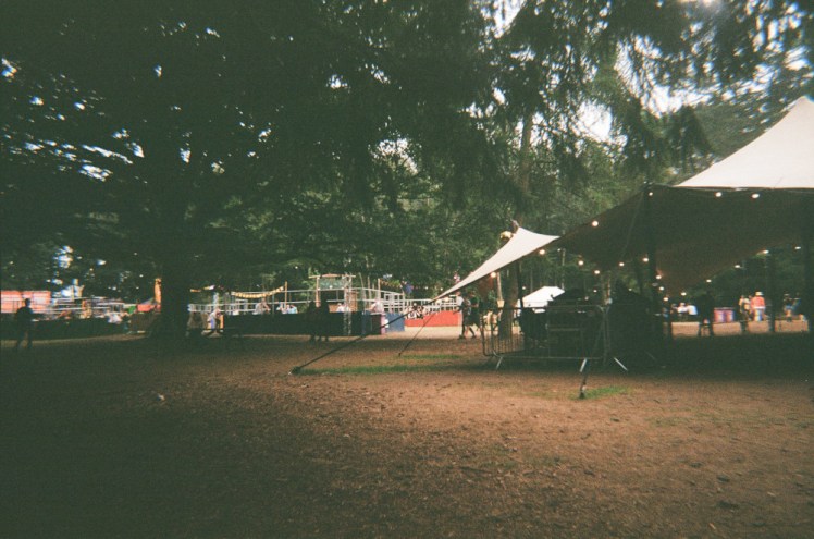 The centre of Camp Wildfire, among cedar trees. There's a tent canopy with a bar and dancefloor disappearing off to the right and to the left in the background is the Patrol Games arena.