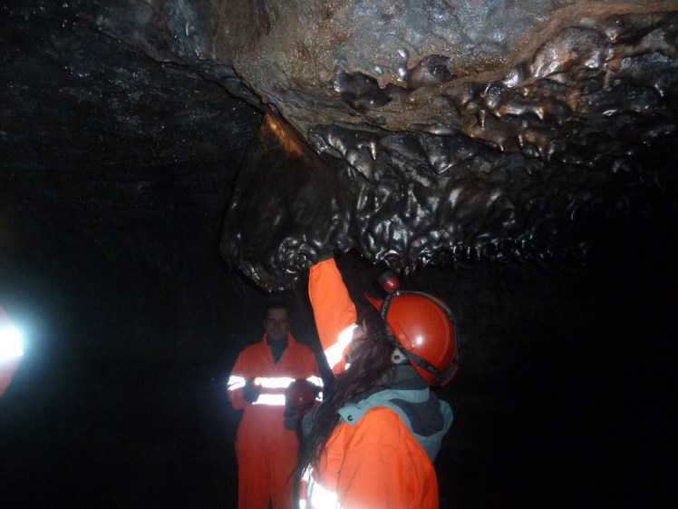 A group, dressed in orange boiler suits with reflective stripes across, reflecting white in the camera's flash, exploring a lava cave. In the foreground, a tourist in a helmet is sticking her hand up into a feature on the low ceiling.
