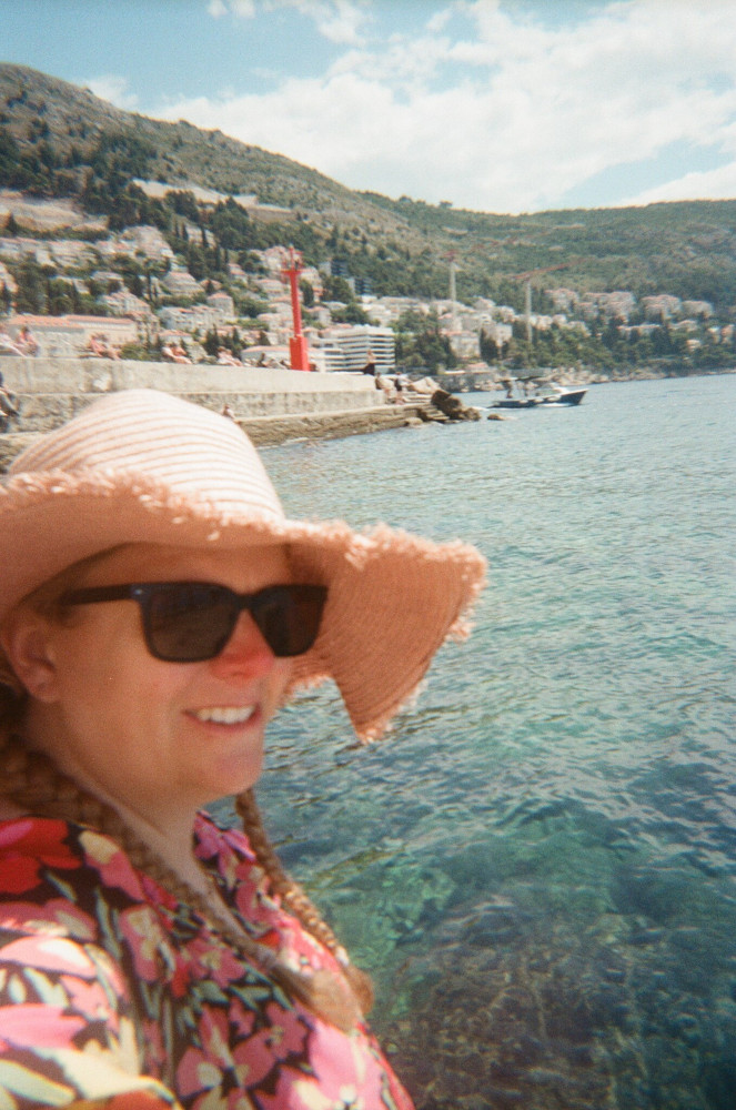 A selfie on the seaward side of Dubrovnik. I'm wearing a pink hat straw hat and a pink flowery dress and the sea behind and below me is vivid turquoise.