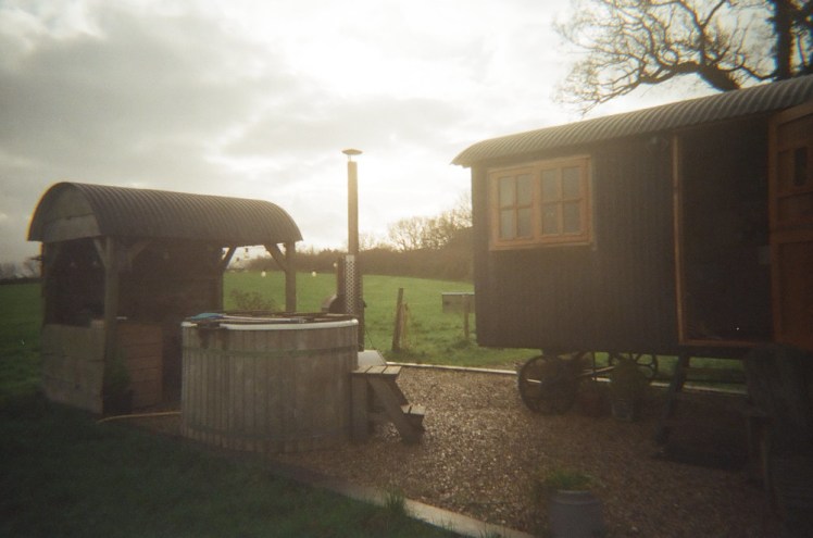 A shepherd's hut, hot tub and dining shelter in shades of greeny-grey, mostly thanks to the sun being quite low and almost behind them.