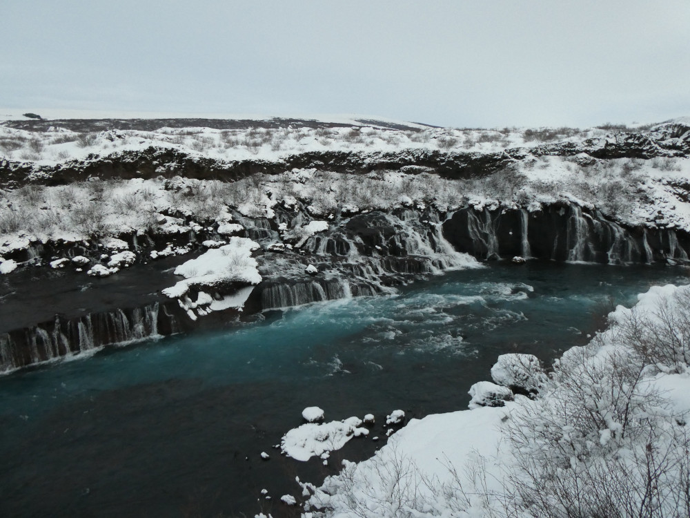 Hraunfossar, a waterfall formed by hundreds of small white rivers of water running down a black lava river bank where there is no river at the top to feed the waterfalls. The picture could almost be in black and white if not for the deep teal-green river flowing along the bottom.