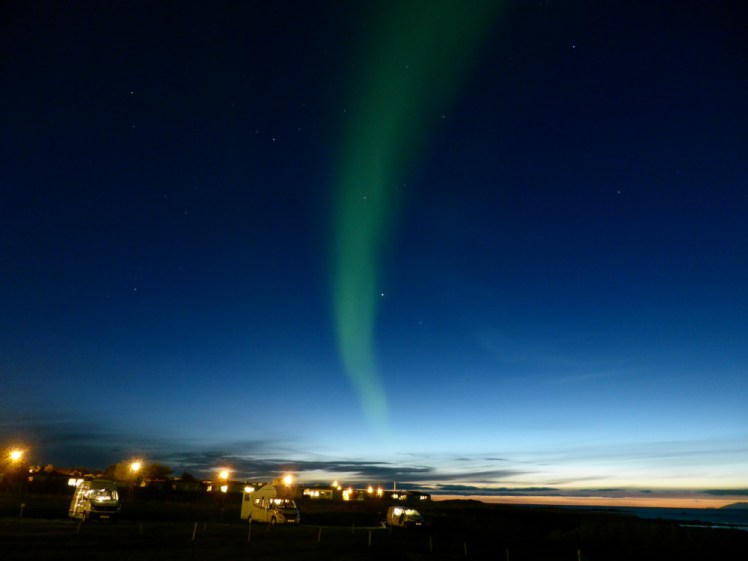 A vertical streak of vivid, unedited Northern Lights over Akranes. The sky is quite light, white-orange sunset just about visible on the horizon, although the highest part of the sky is deepest navy blue.