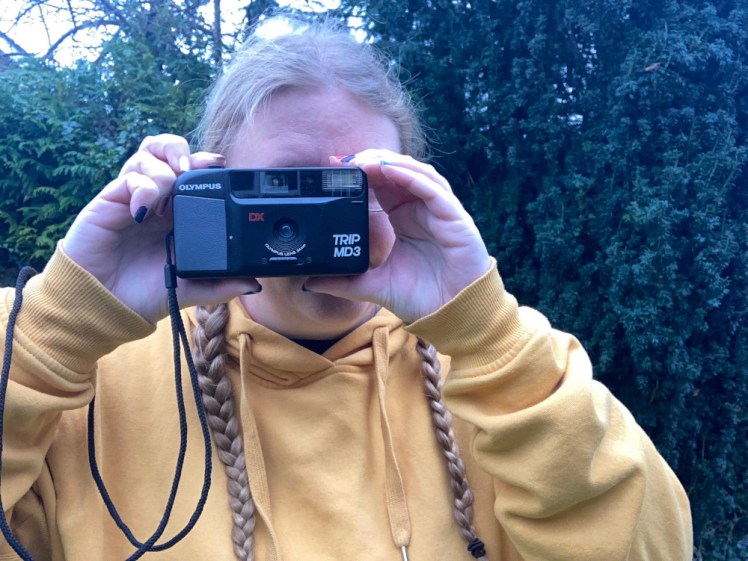 Me in a yellow hoodie holding up a black 90s point-and-shoot film camera. The picture was taken on my phone yesterday and yet has a weird blueish tinge that almost makes it look like it was also taken on film.