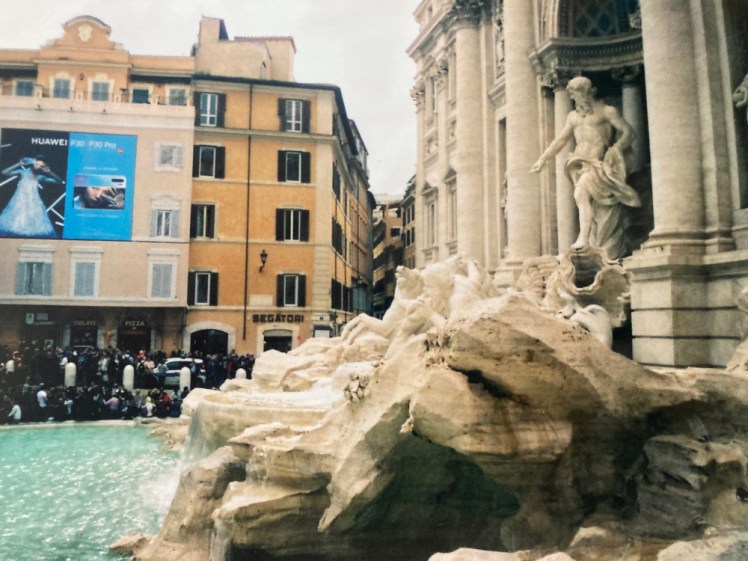 The Trevi Fountain as seen from the side. On the opposite side of the pool, you can see hundreds of people crammed in together taking photos of it.