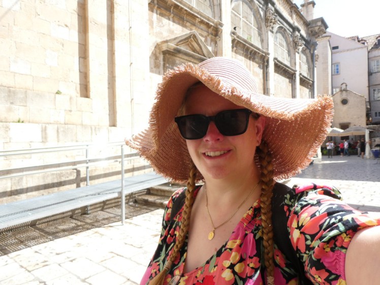 A selfie in the streets of Dubrovnik. I'm wearing a short dress covered in big bold pink flowers and an enormous pink straw hat with a floppy brim.