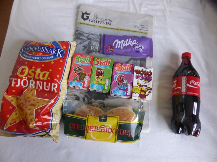 A little haul of supermarket snacks including some cheese-flavoured star-shaped crisps, hamburger rolls, cartons of juice & chocolate milk, a bar of chocolate, a small bottle of Coca Cola, all lying on top of a Reykjavik Grapevine.