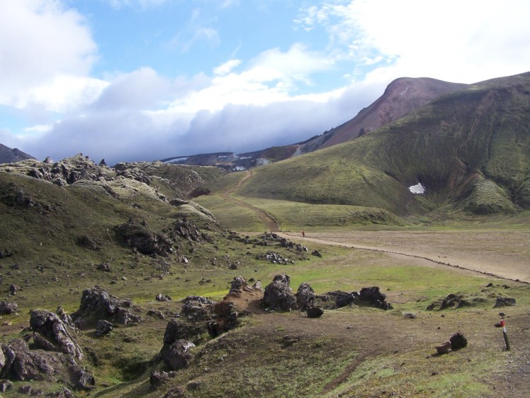 A dirt track running across a moss-green field and up and over moss-green hills towards the mountains in the distance.