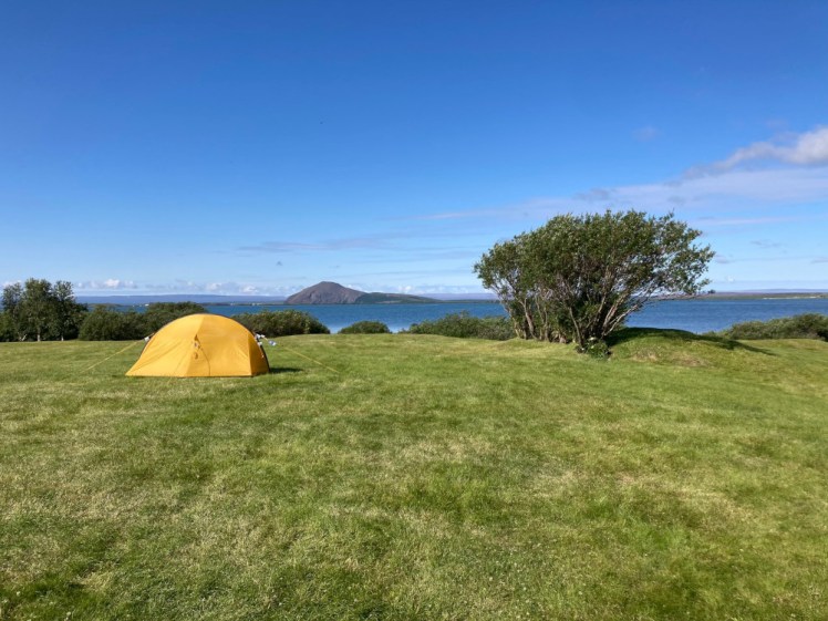 A tiny little yellow one-man tent in an off-green field with a storm-blasted tree on the edge of a blue lake under a blue sky. There is a nice triangular mountain on the other side of the lake.