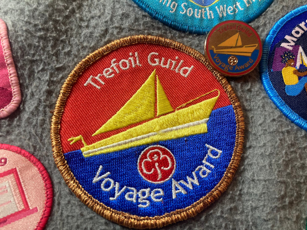 Bronze Voyage Award badges - a large red and blue badge with a yellow boat on it and a bronze metallic border is sewn on my grey fleece blanket. Next to it lies a smaller metal badge in much the same design except the sky is bronze and the rim is red.