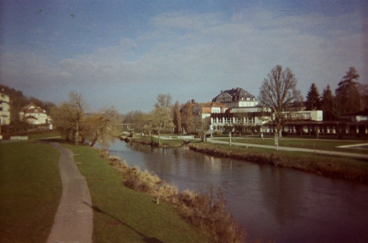 The middle of Bad Kissingen as seen from the bridge. There's a river flowing underneath with grassy park on both sides and some villas above the grass.